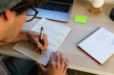 high school or college student with glasses studying and writing on a desk with a laptop from top...