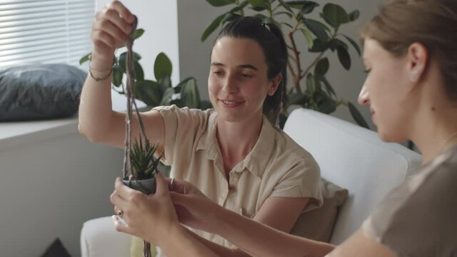 Two young cheerful women putting flowerpot into handmade macrame plant hanger and discussing it while sitting at home