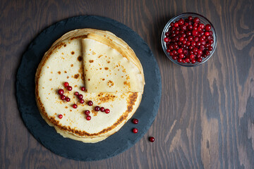 Top view of pile or stack of russian homemade traditional blinis or crepes with red bilberry berries served on round stone tray on dark brown wooden background. Image with copy space, horizontal