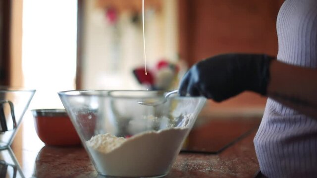 Detalle de una mujer con guantes negros vertiendo aceite de oliva en una cuchara y un recipiente de vidrio transparente con harina en la encimera de la cocina.