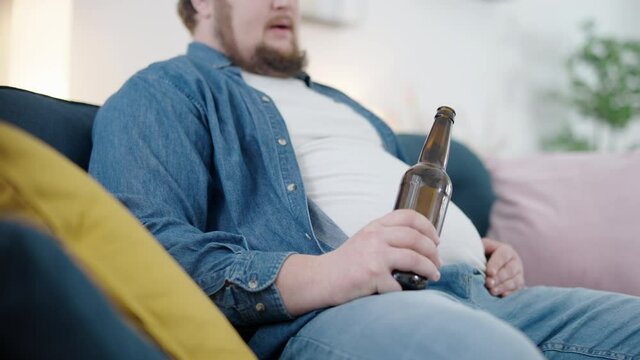 Relaxing bachelor drinking beer from bottle rubbing fat belly, weight gain, rest