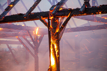 Burned down wooden barn with fire and sparks still blazing. Charred roof truss and wooden posts. - obrazy, fototapety, plakaty