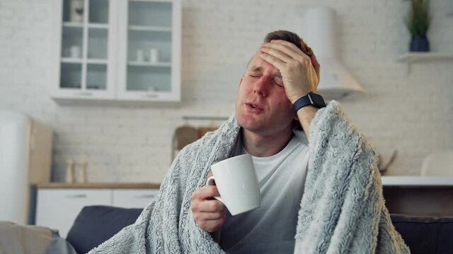 Folk medicine. Sick young man with fever wrapped in warm blanket drinking hot tea. Pandemic.