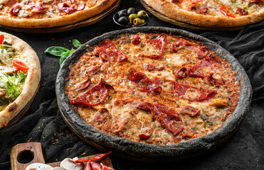 Pizza Pepperoni with black dough, prosciutto, sausage, cheese parmesan, spinach on stone black background. Fast food lunch for picnic company, top view, selective focus