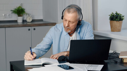 An elderly man wearing headphones sits at a laptop and makes notes.