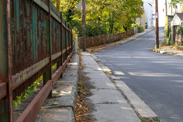 Rusting metal fence alongside a very narrow sidewalk, road, yellow leaves of early fall, horizontal aspect