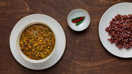 Whole mung bean curry with red rice and chili on wooden table.