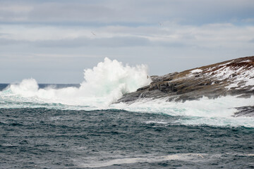 A rugged coastline on a sunny day with blue sky and clouds. The ocean is raging and is hitting the side of the craggy cliff. There's the land in the background and the teal ocean in the foreground.