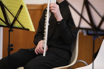 Flute musical instrument of silver color in an upright position in hands against the background of a black shirt and trousers of a seated man with music and music stand.Concept of a school concert