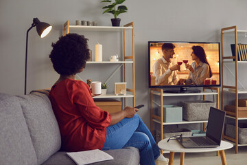 Woman enjoying good romcom serial in the evening. Single African American lady sitting alone at home, drinking coffee and watching romantic love story on soap opera entertainment TV channel, back view