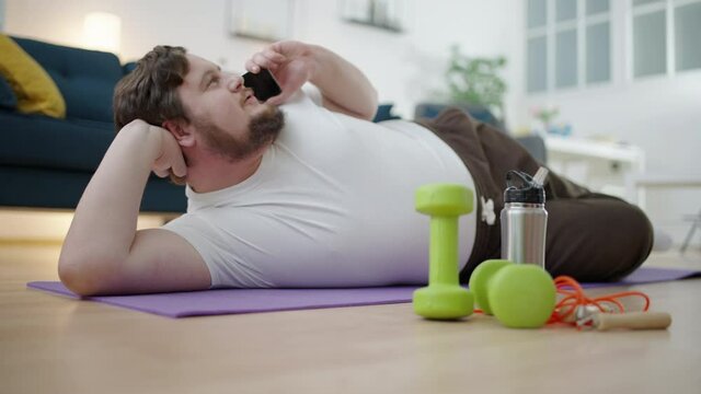 Overweight man lying on exercise mat in front of dumbbells, talking on phone