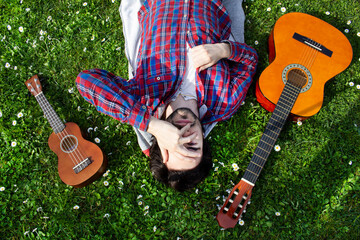 Young man lying on the grass field with flowers around, acoustic guitar and ukulele around him