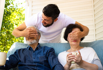 Happy brunette adult son standing behind senior parents, covering them eyes and making surprise, while mature father and mother sitting on sofa at home terrace outdoors. Generation and people concept