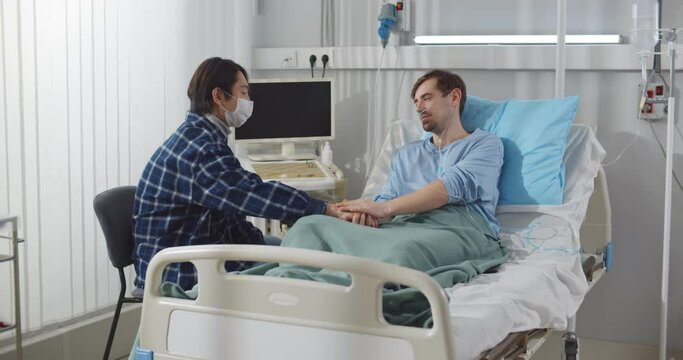 Young man in protective mask holding sick friend hand while sitting by bed in hospital