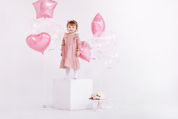 happy birthday 2 years old little girl in pink dress. white cake with candles and roses. Birthday decorations with white and pink color balloons and confetti for party on a white wall. Happy birthday