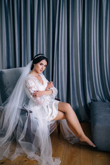 Fototapeta premium A serious, excited beautiful bride sits on a gray armchair by the window. morning before the wedding.