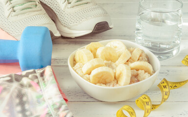 oatmeal with banana slices, glass of water, blue dumbbells, sneakers, sportswear, measuring tape on a white