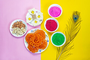 Colorful holi powder or gulal in ceramic bowl along with traditional Indian sweet food imarti, sandesh, cashew and almonds in plate on multicolor background.