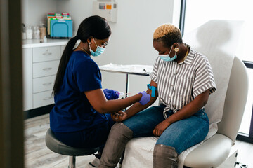 Black nurse collects blood sample from black female patient in exam room at medical facility