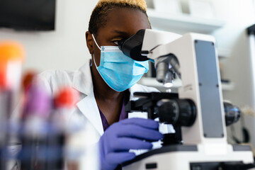 Black medical technologist in mask in laboratory looking through microscope at blood samples