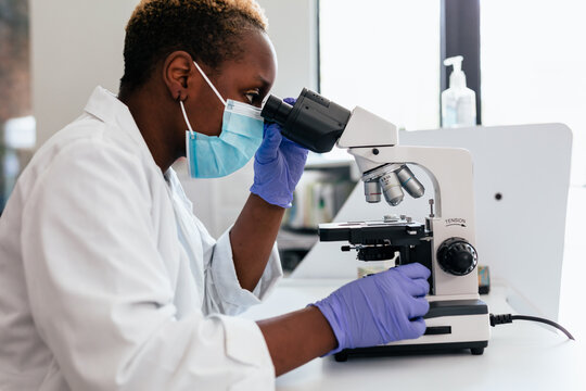 Black female scientist in mask in laboratory looking through microscope