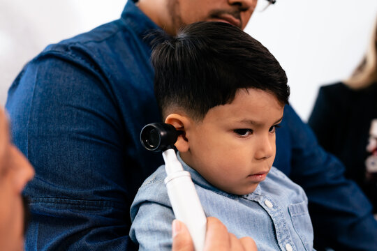 Doctor Exams Ears Of Young Hispanic Toddler With Otoscope In Medical Exam Room During Wellness Visit