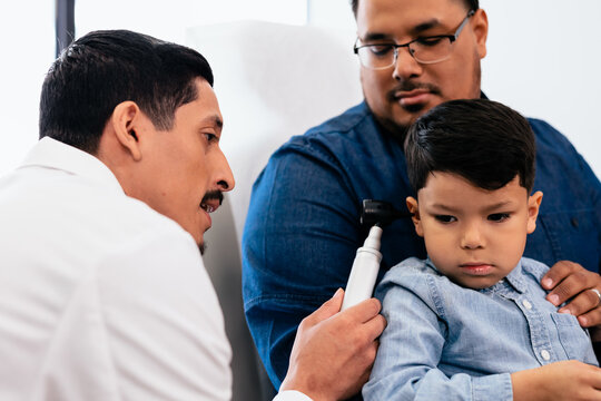 Doctor Exams Ears Of Young Hispanic Toddler With Otoscope In Medical Exam Room During Wellness Visit