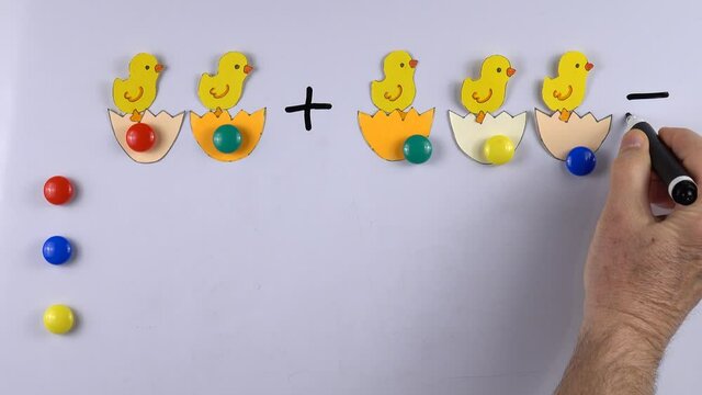 Teacher teaches kids basic of math addition with the aid of magnets and figures of chicks, cut out from paper, on the whiteboard, example of educational math children game