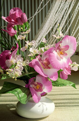 Home decoration floral display of artificial pink and white flowers in white round vase. White net curtain and dark green stripe wallpaper background. Sunlight from window making patterns.