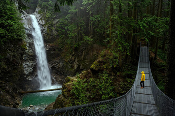 Woman in yellow rain jacket standing on a suspension bridge and looking at the Cascade falls, in Cascade falls regional park, Deroche, British Columbia, Canada