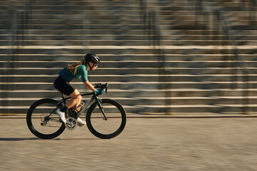Side view of professional female cyclist in cycling garment and protective gear riding bicycle in city, rushing and passing buildings while training outdoors on a daytime - obrazy, fototapety, plakaty