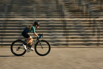 Plexiglas foto achterwand Side view of professional female cyclist in cycling garment and protective gear riding bicycle in city, rushing and passing buildings while training outdoors on a daytime © Friends Stock