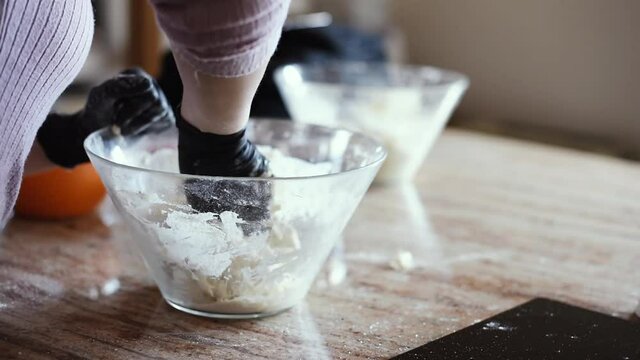 Una mujer en la cocina amasando una masa de pizza dentro de un bol con harina mientras usa guantes negros.