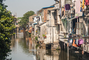 Fototapeta na wymiar Suzhou China - May 3, 2010: City canal with brown water reflects the backside of houses along the water, and green foliage on other side under gray sky. Lots of laundry adds colors.