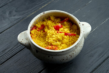Appetizing and dietary oriental side dish - couscous porridge with vegetables served in a white pot on a black wooden background.
