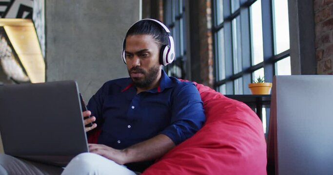Happy mixed race man sitting in cafe listening to music on headphones using smartphone and laptop