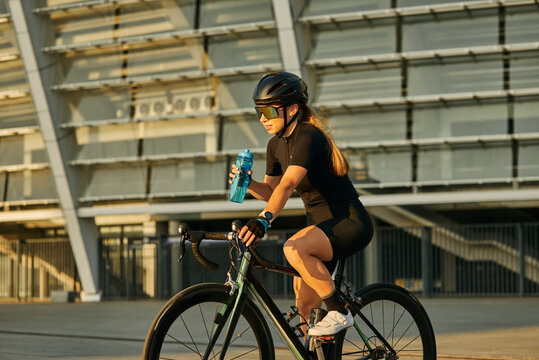 Professional female cyclist in black cycling garment and protective gear holding water bottle while riding bicycle in city, training outdoors at sunset