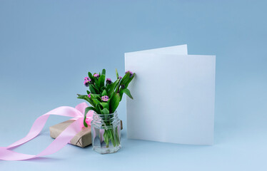 Blank greeting card with small bouquet of forget-me-not flowers and small present box with pink ribbon on light blue background