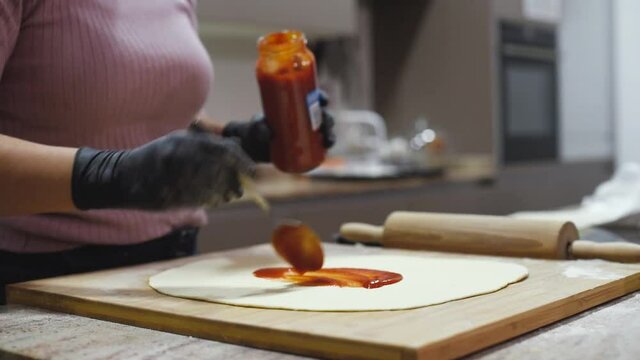 Una mujer con guantes extendiendo salsa de tomate con una cuchara sobre una base de pizza casera. Bote de salsa de tomate.
