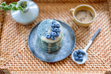 Rattan tray with blue american pancakes with blueberry poured with caramel souse, cup of coffee and vase with flower. Homemade tasty food. Celebration of Shrovetide.