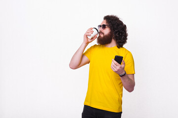 Photo of bearded hipster man in yellow t-shirt drinking coffee and holding smartphone