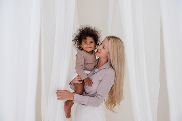 Young blond Caucasian mother is holding African American curly daughter in arms. Woman hugs girl kisses among the white cuts of floating light tulles. Mother's love care. Adoption, mixed ethnic family