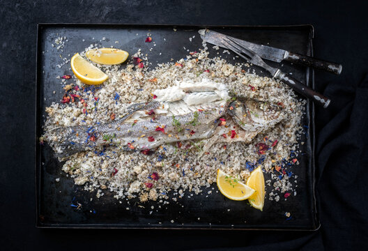 Traditional Atlantic Loup de Mer bedded into sea salt served with lemon slices and spices as top view on a rustic metal tray