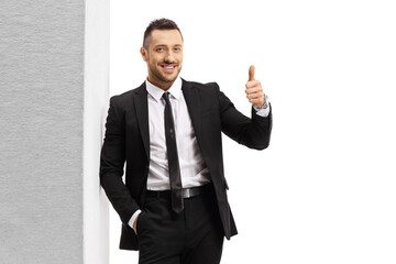 Nan in a black suit and tie leaning on wall and showing thumbs up