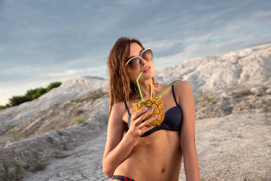 Summer heat. A girl in a bathing suit and sunglasses with a cocktail and pineapple in their hands drinking fresh juice from a straw against the background of white hills and cracked clay.