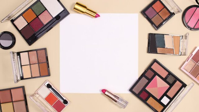 Eye shadow make up palettes and lipsticks appear around paper card note on nude background. Stop motion flat lay
