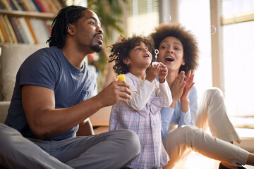 Young parents having a good time with their cute little daughter at home while making soap bubbles together. Family, home, playtime