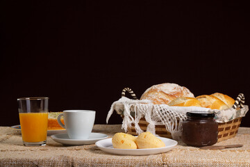 Table set for breakfast or coffee break, with a basket of breads, jam, cake, cup of coffee, glass of orange juice and chipas or pão de queijo or Brazilian cheese bread, on brown tablecloth.