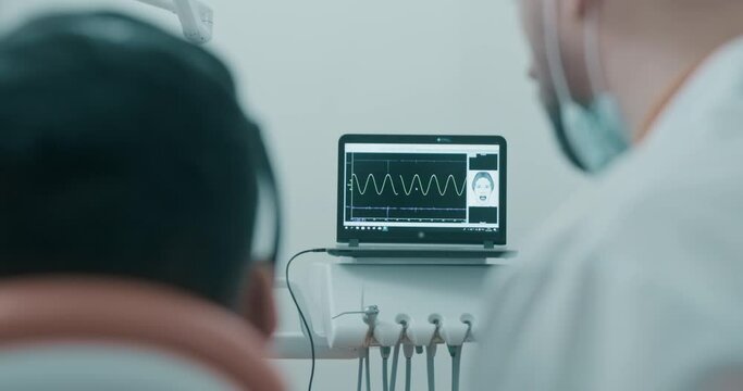 A male patient having a Joint vibration analysis procedure (JVA) and laying in the dental chair while male dentist standing near to him and they are looking to the screen with a line of vibration.