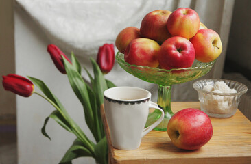 Still life, consisting of a bouquet of red tulips in a jug, a bowl of apples, a cup of tea and a bowl of coconut sweets.